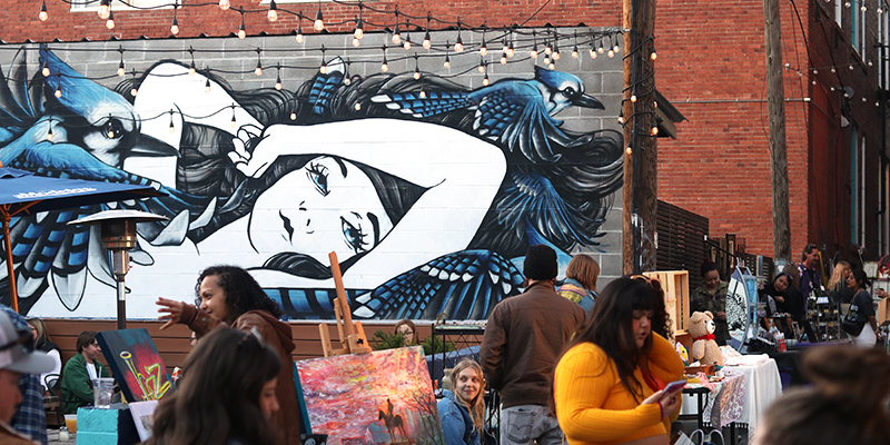 Blue and white mural of woman resting with her arm on her head, people walking through an alley next to the mural